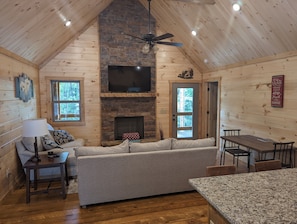 Relax by the stone fireplace in this cozy cabin-style room, featuring a comfortable sofa and dining area with a view of the surrounding forest.