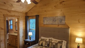 Ein Schlafzimmer einer Holzhütte mit Bett, Nachttisch und Fenster mit Blick auf Bäume.