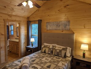 A wooden cabin bedroom with a bed, nightstand, and a window with a view of trees.