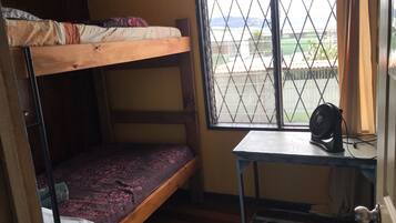 Un dormitorio con literas, una mesita con un ventilador y una ventana que da a vistas del exterior.