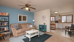 A living room with a beige sofa, a coffee table, a bookshelf, a ceiling fan, and a kitchen area with a microwave. - Are you traveling to Rochester? Be our guest!!  (Rochester)