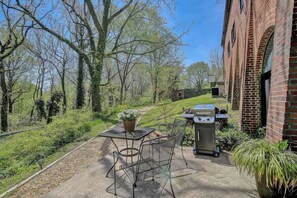An outdoor patio with a grill, a table, and chairs, surrounded by trees and a brick building.