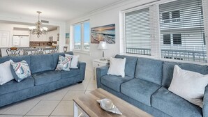 A modern living room with two blue sofas, a wooden coffee table, and a view of the city through large windows. - Hidden Treasure -3BR Sleeps 8 (Navarre Beach)
