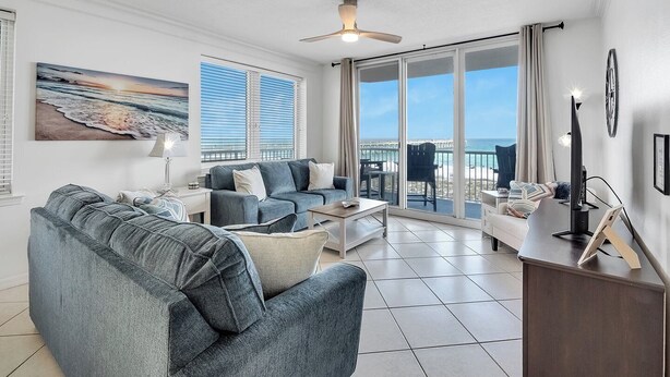 A spacious living room with a large gray sofa, a coffee table, and a TV stand. There is a balcony with a view of the ocean. - Hidden Treasure -3BR Sleeps 8 (Navarre Beach)