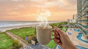 Une main tenant une tasse de laquelle s'échappe de la vapeur, avec en arrière-plan une plage et un espace piscine bordé de bâtiments.