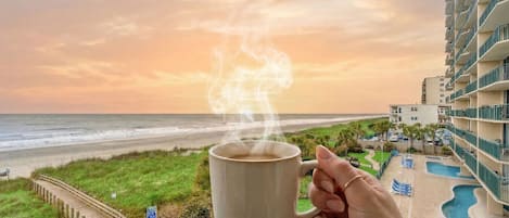 Una mano sostiene una taza con vapor, con vista a una playa y una piscina, con edificios al fondo.