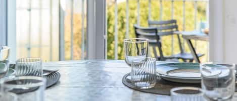 Et spisebord dækket med tallerkener, glas og bestik, med udsigt til en balkon og udendørs siddepladser.