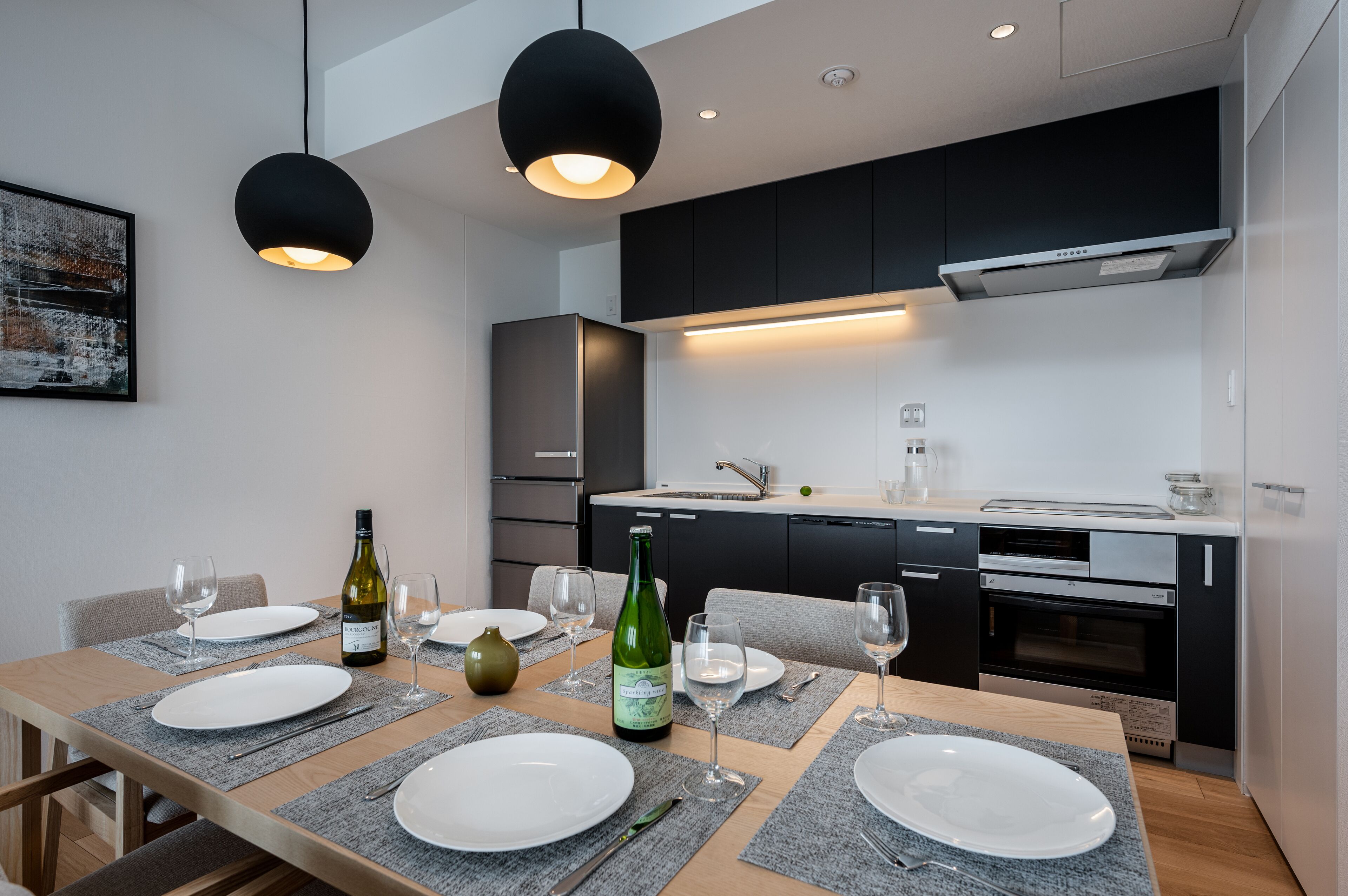 A modern kitchen with black cabinets, a built-in oven, and a dining area set with white plates and wine glasses.