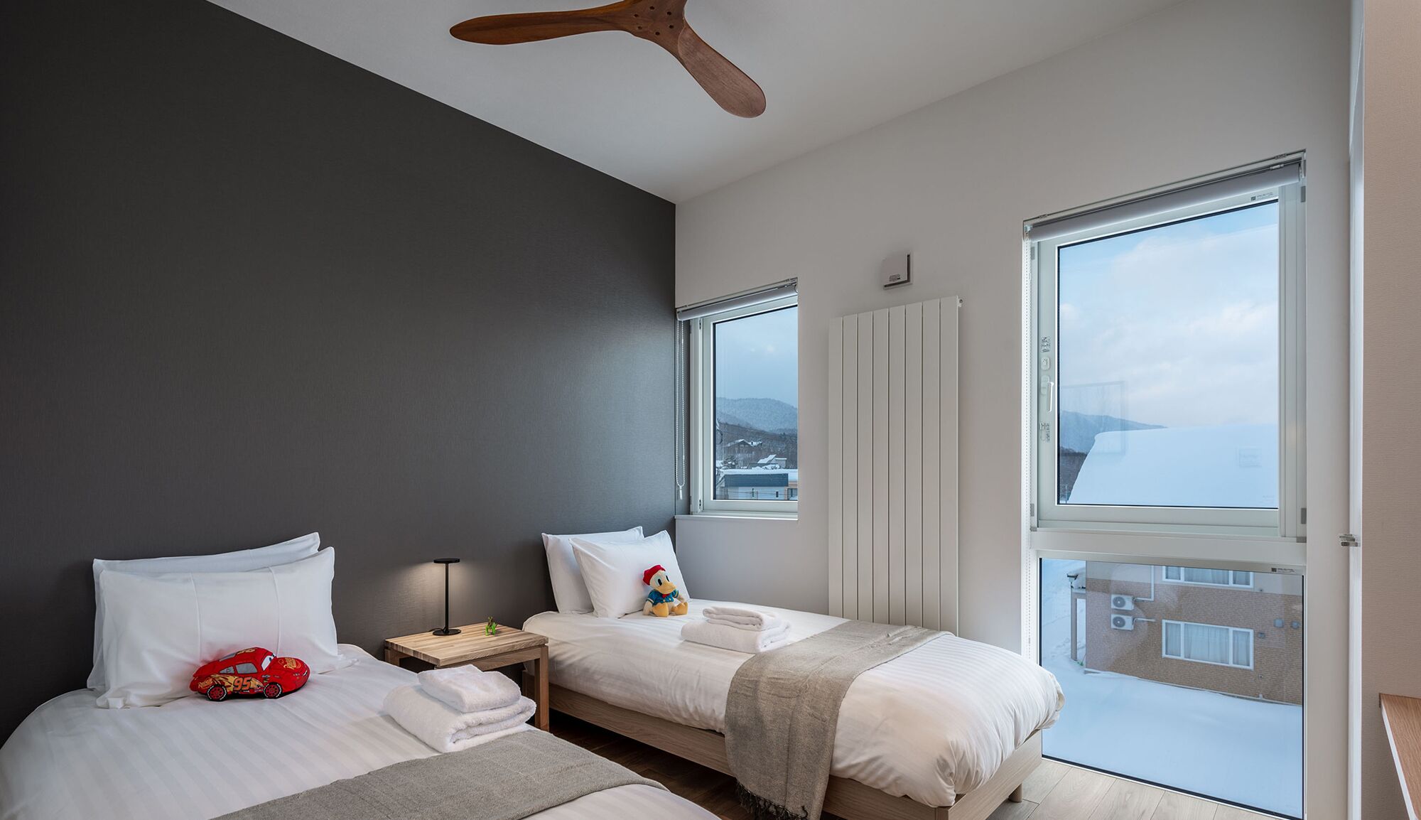 Quarto de hotel moderno com duas camas, um ventilador de teto de madeira, mesa de cabeceira, luminária e amplas janelas com vista para o mar.