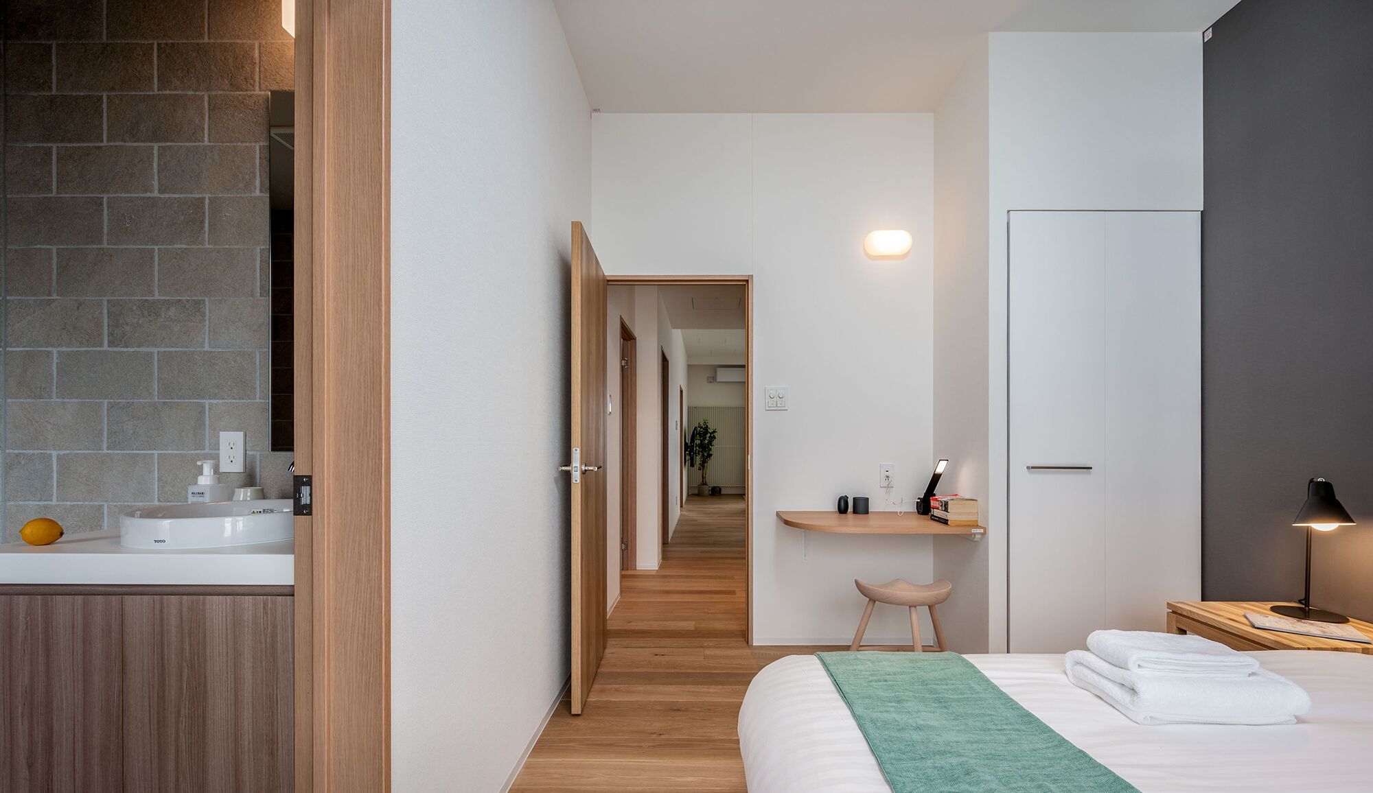 Quarto de hotel moderno com piso de madeira, cama com manta verde, mesa de cabeceira com abajur, escrivaninha com cadeira e banheiro com pia e espelho.