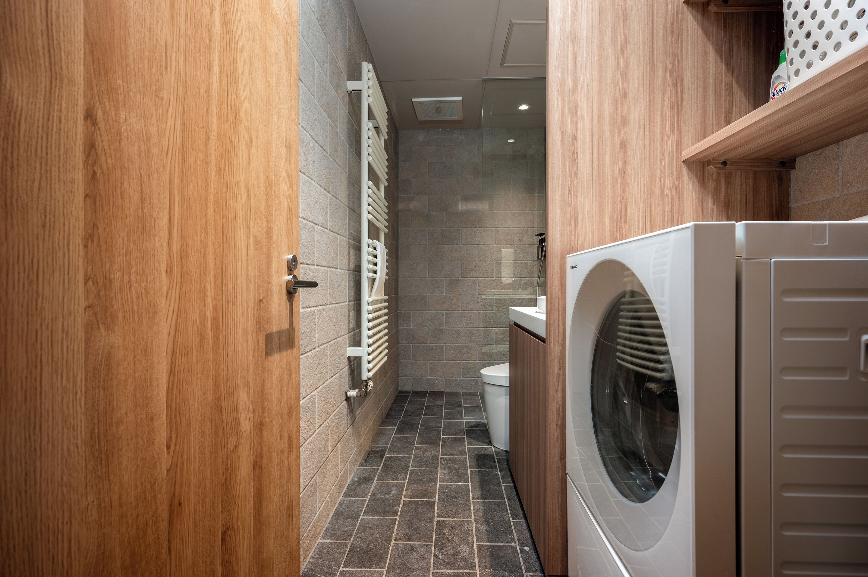 A compact bathroom with a wooden door, a built-in shower, and a front-loading washing machine.