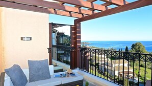 Uma varanda com vista para o mar, um sofá com almofadas, uma mesa pequena com frutas e um livro.