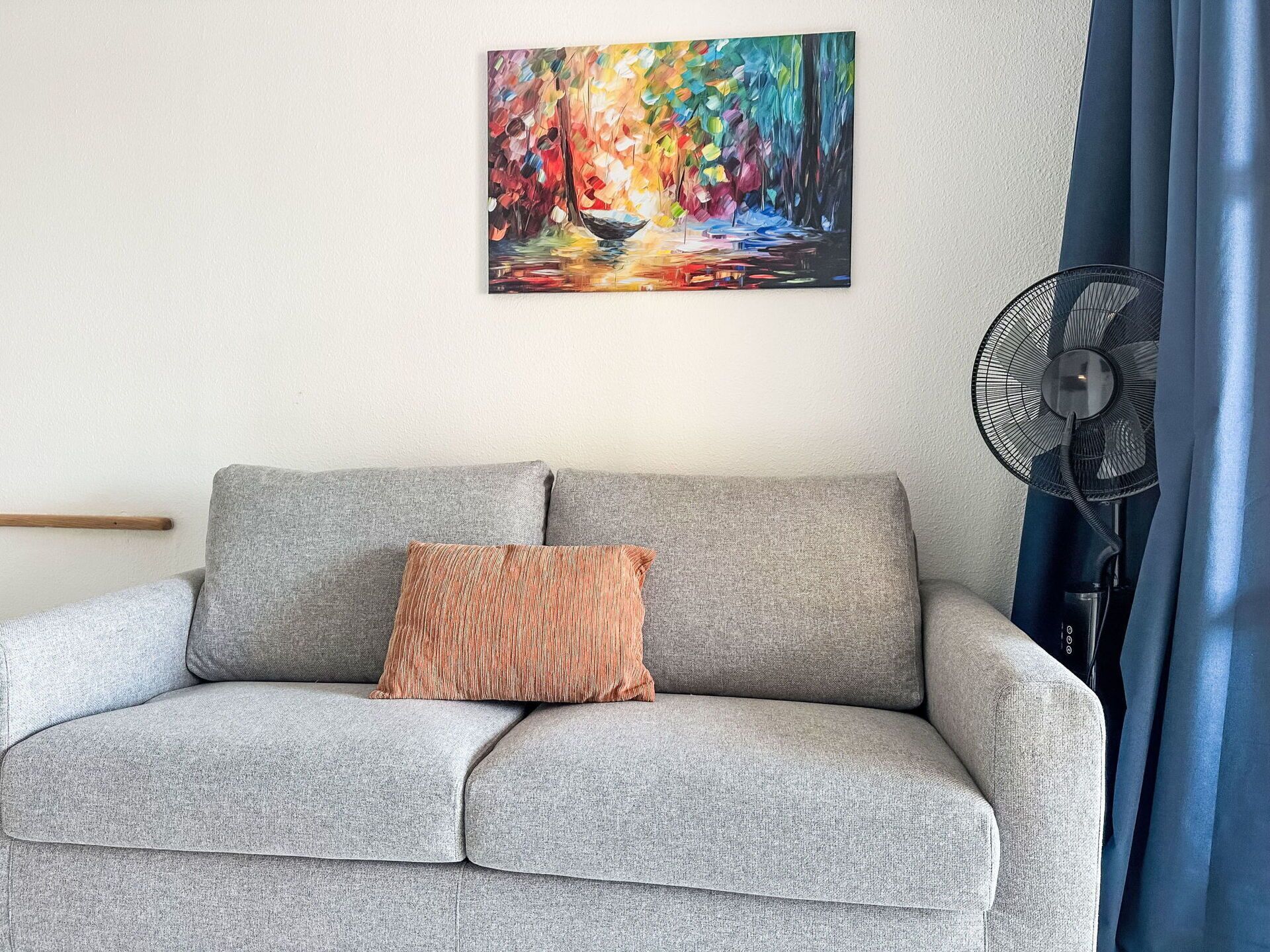 Ein graues Sofa mit einem einzelnen gestreiften Kissen, einem an der Wand hängenden Bild und einem Standventilator.