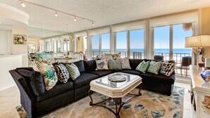 A spacious living room with a large black sectional sofa, a glass-top coffee table, and a view of the ocean through large windows. - Oceanfront | Turtle Dunes Luxury Penthouse | Intracoastal and Oceanfront Views | Double Balconies (Fernandina Beach)
