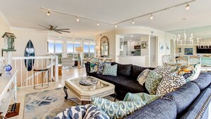 A spacious living room with a dark-colored sofa, a glass coffee table, and a white railing. There is a ceiling fan, a chandelier, and a large mirror. - Oceanfront | Turtle Dunes Luxury Penthouse | Intracoastal and Oceanfront Views | Double Balconies (Fernandina Beach)