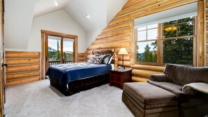 Ein Schlafzimmer in einer Blockhütte mit einem Bett, einem Nachttisch, einem Sessel und großen Fenstern mit Blick auf die Berge.