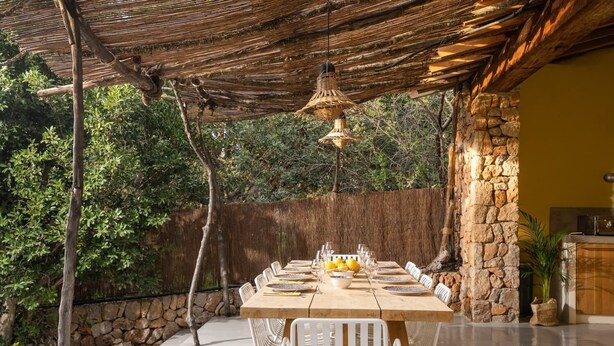 An outdoor dining area with a stone wall, a wooden table set with white chairs, and a hanging light fixture. - Ca'n Quinn (Deià)
