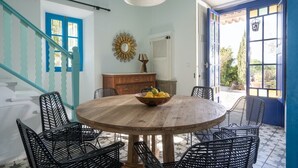 A round wooden table with a bowl of fruit, surrounded by black metal chairs, in a room with teal walls, a white ceiling, and a blue door. - Ca'n Quinn (Deià)