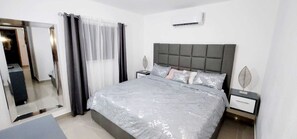 Une chambre à coucher moderne avec un grand lit, une literie grise, une tête de lit grise et une table de chevet agrémentée d’un objet décoratif.