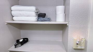 A bathroom with white shelves holding towels and a toilet paper roll. There is a power outlet with a hairdryer plugged in.