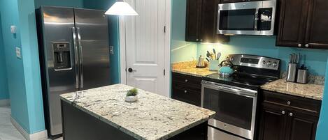 Una cocina con isla central, electrodomésticos de acero inoxidable, armarios de madera oscura y una pared verde azulado.