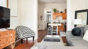 Uma sala de estar moderna com televisão, uma poltrona estampada, um sofá cinza e uma área de cozinha com armários de madeira e um banco de bar.