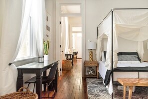 Un dormitorio con cama con dosel, una mesita de noche con lámpara, un pequeño ventilador y una planta en maceta.
