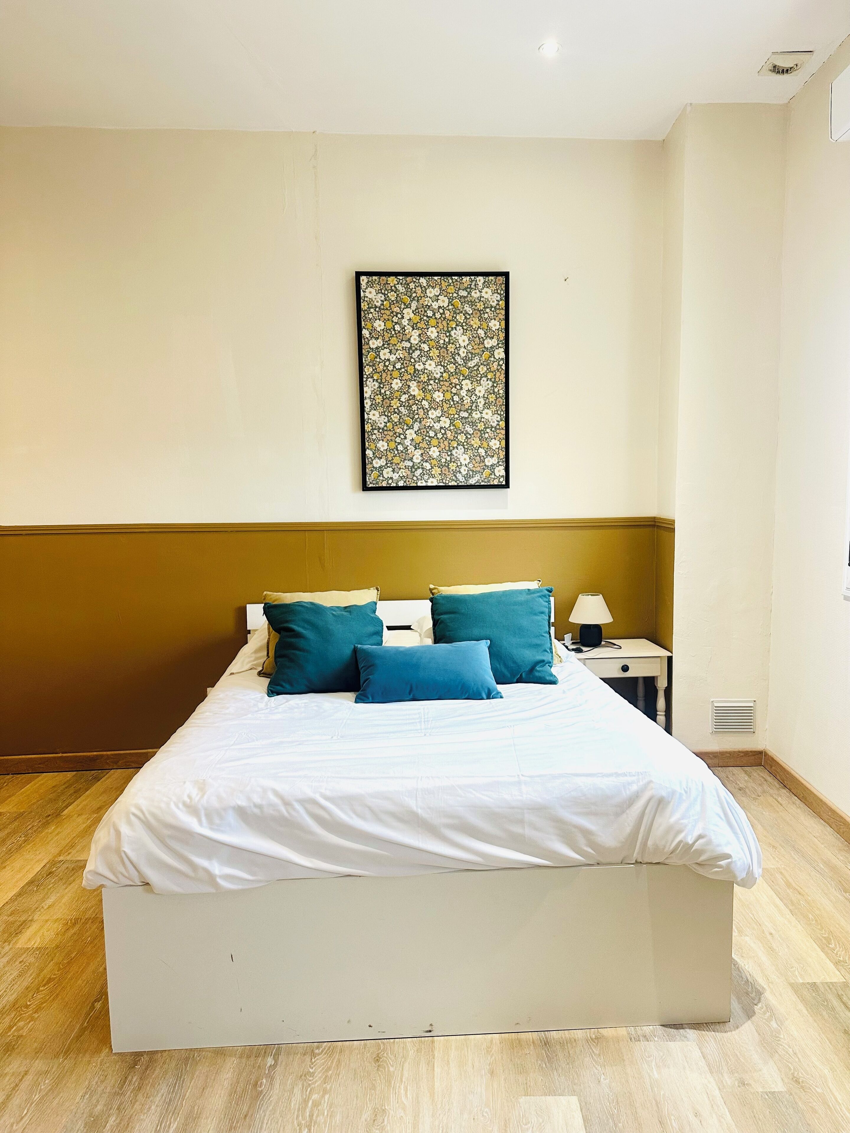 Ein ordentlich bezogenes Bett mit weißen Leinen und türkisfarbenen Kissen, ein kleiner Nachttisch mit Lampe und ein Blumenbild an der Wand.