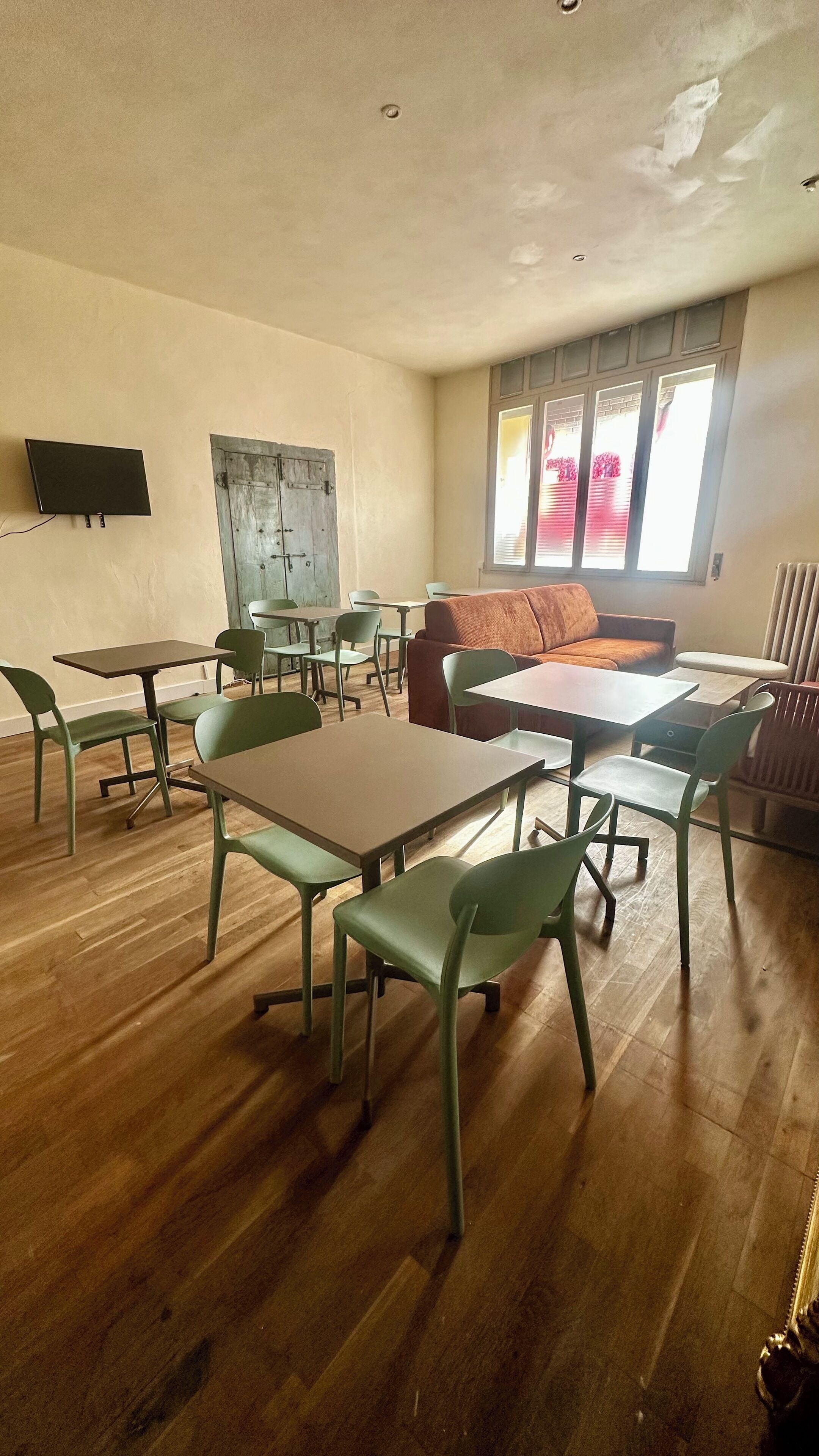 Habitación con suelo de madera, sillas verdes y mesas dispuestas para comer. Hay un sofá y un radiador cerca de la ventana.
