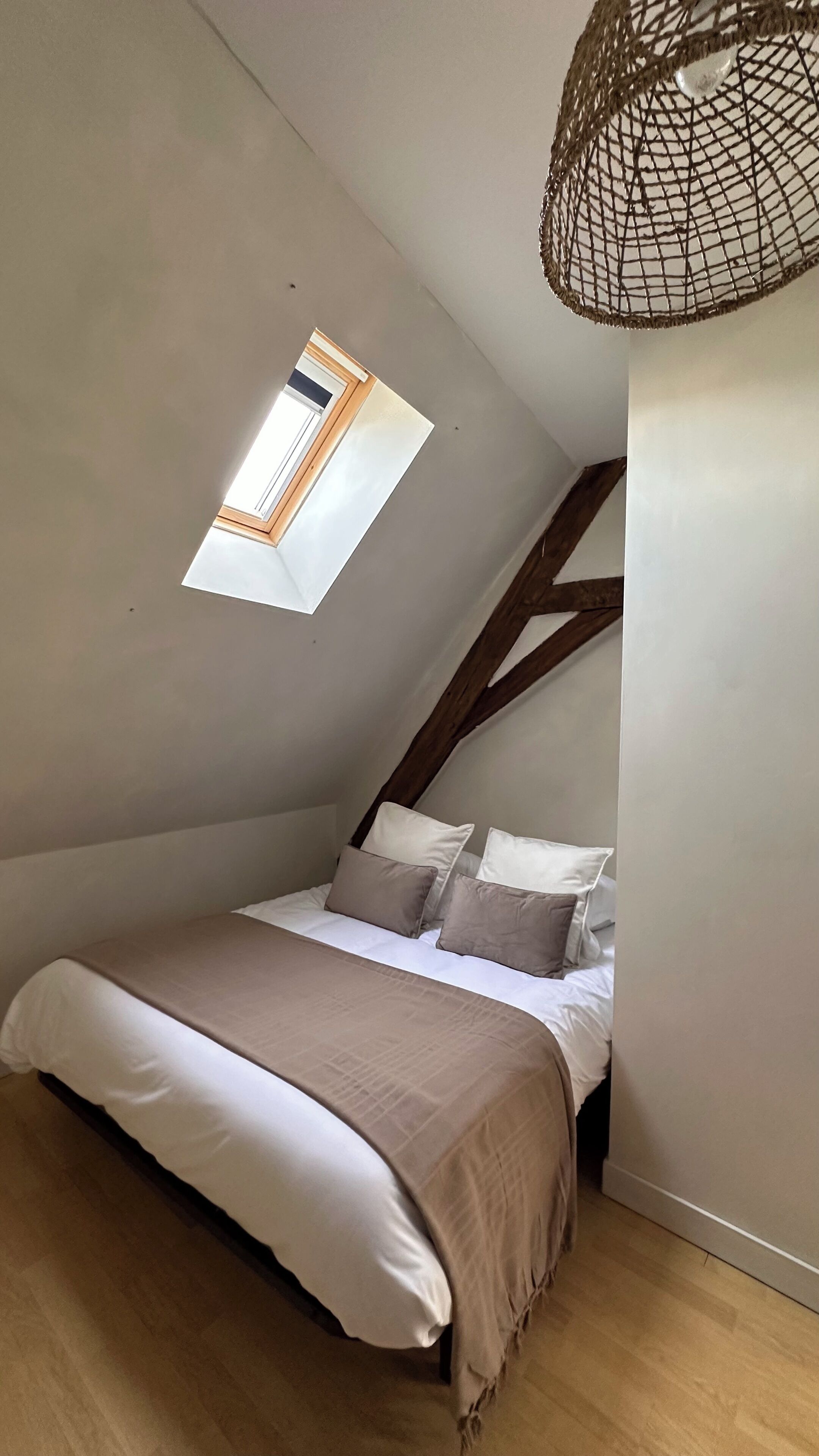 Une chambre avec un plafond en pente, une lucarne, un lit avec une literie blanche et beige, et une poutre en bois au plafond.