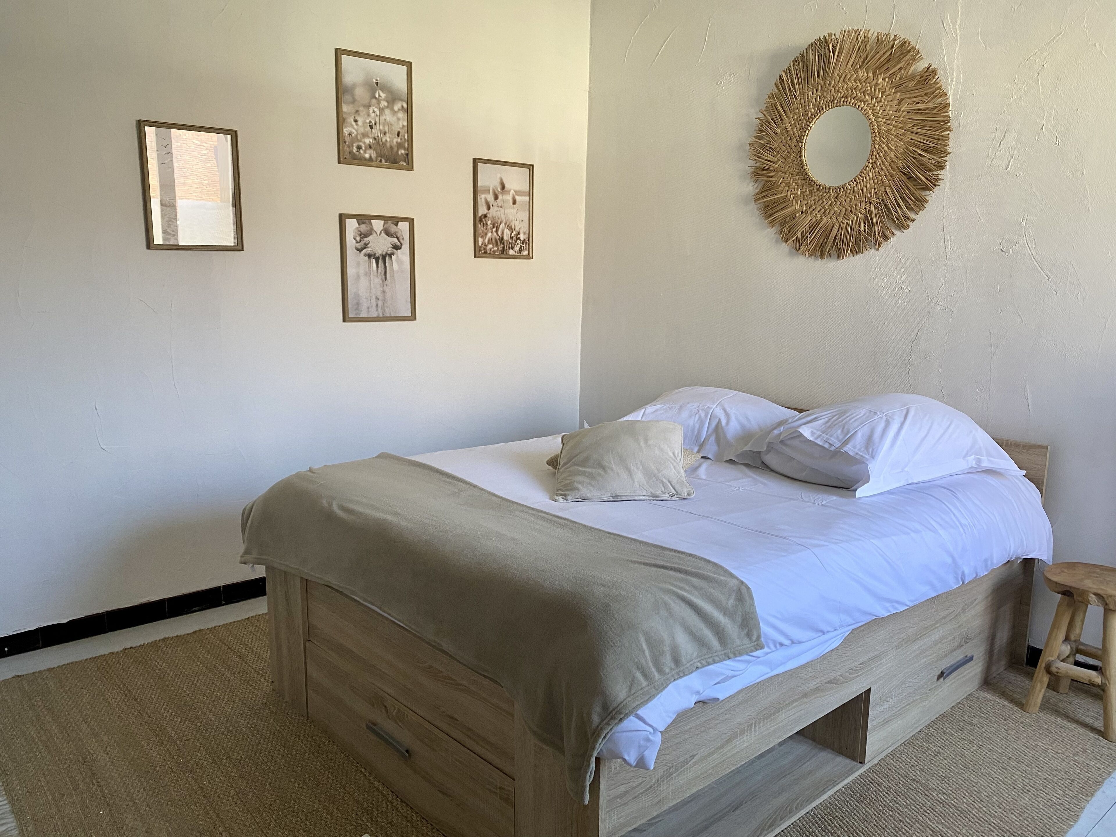 Een slaapkamer met een houten bed, een beige deken en een houten krukje. Op de muur hangen vier ingekaderde foto’s.