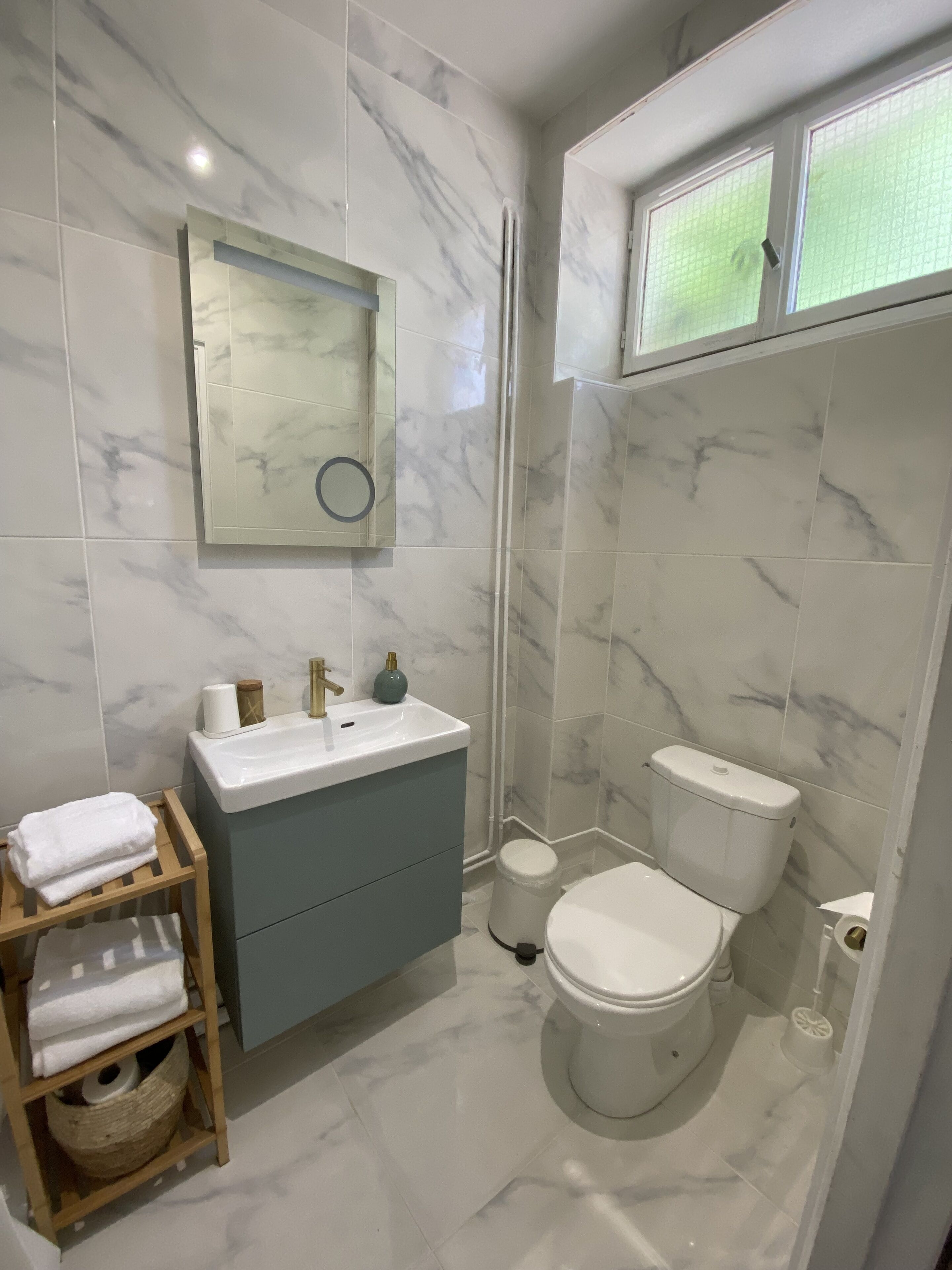 Une salle de bain moderne avec un pare-baignoire en marbre, un lavabo avec un miroir au-dessus, des toilettes et une petite fenêtre.