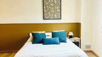 A neatly made bed with white linens and blue pillows, a small bedside table with a lamp, and a floral artwork on the wall.