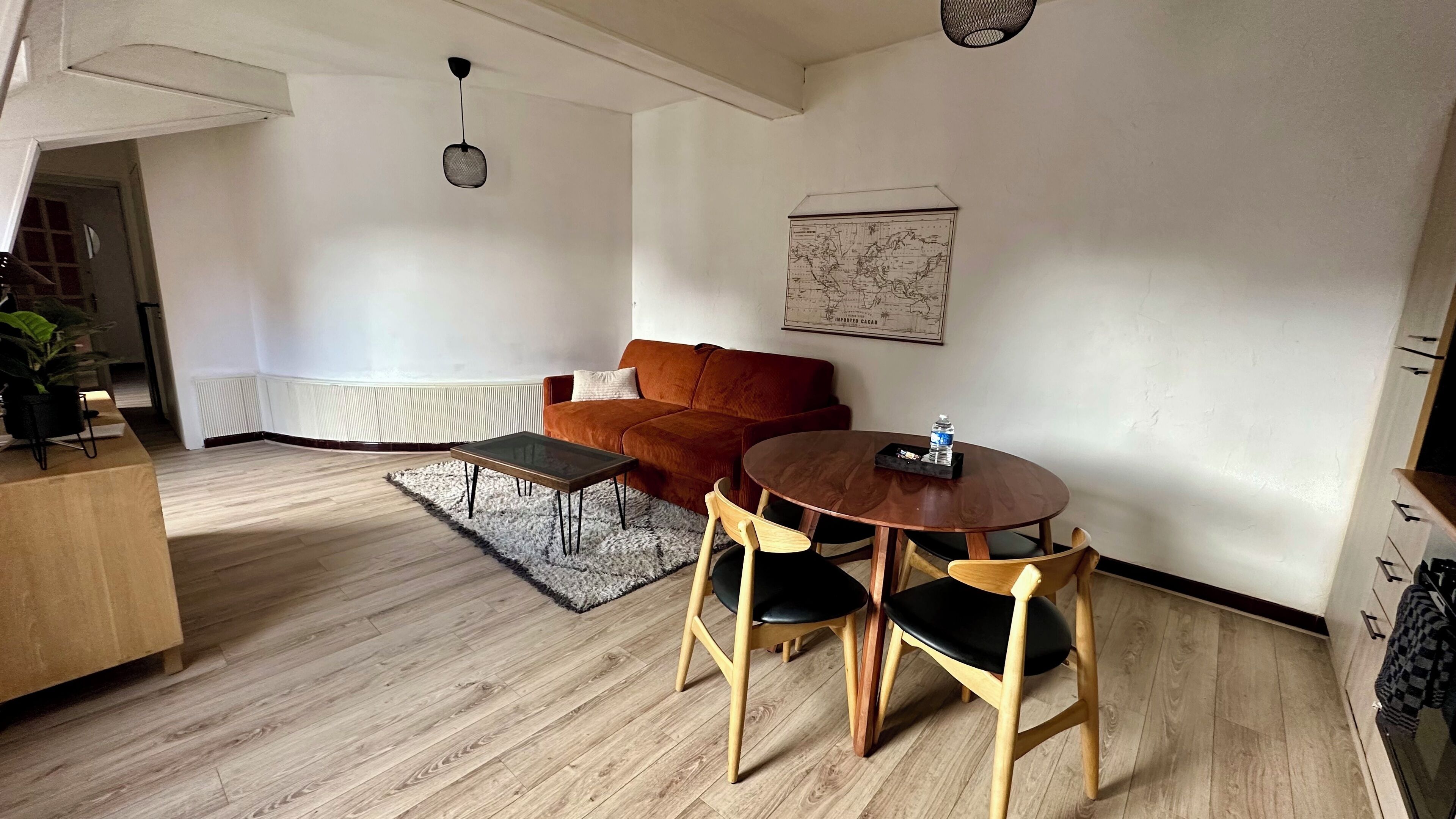 A modern living room with a wooden floor, a brown sofa, a coffee table, and a dining table with chairs. There is a map on the wall and a ceiling fan.