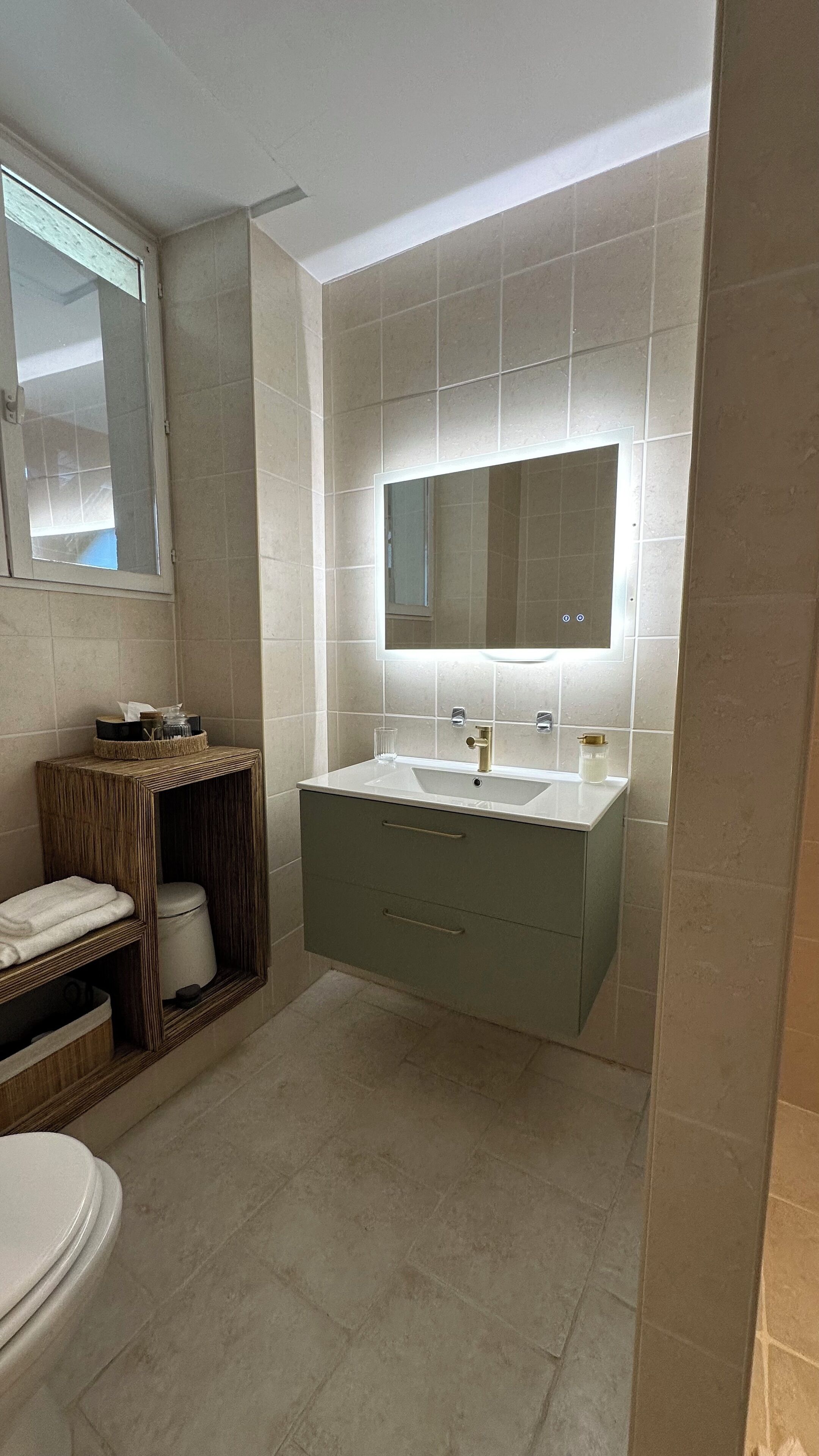 A bathroom with a white sink, a mirror, and a toilet. There is a small shelf with a jar and a tissue box.