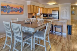A dining area with a wooden table and white chairs, a kitchen with wooden cabinets and stainless steel appliances, and a painting of a harbor scene on the wall.