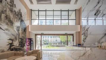 A spacious lobby with a high ceiling, a large painting on the wall, and a view of the outside through large windows.