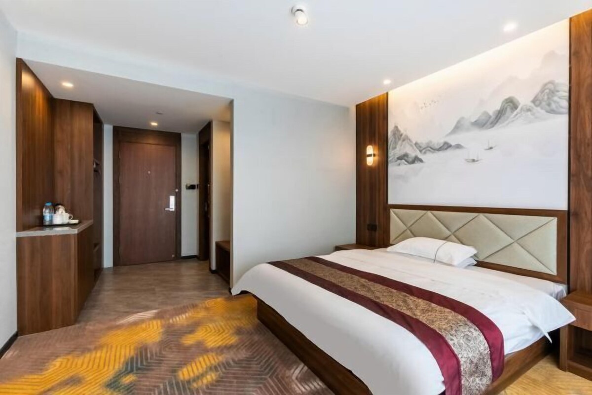 Une chambre d’hôtel avec un lit, une tête de lit en bois, un mur orné d’un tableau représentant une montagne et une porte donnant sur une autre pièce.
