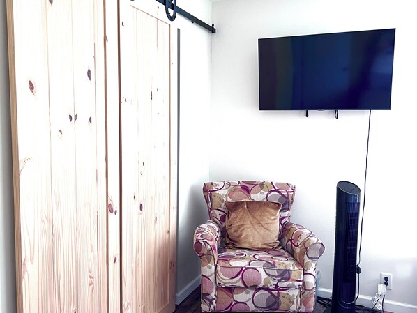 Ein Zimmer mit einer Holztür, einem gemusterten Sessel mit Kissen sowie einem an der Wand befestigten Fernseher.