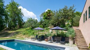 A rectangular swimming pool with a stone border, surrounded by a grassy area and trees. There are lounge chairs and umbrellas under a wooden deck. - La Rocchetta 1915 by Interhome (Cortazzone)