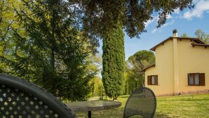 A patio with a round table and two chairs, a tall tree, and a yellow building with windows. - Villa Le Pine by Interhome (Anghiari)