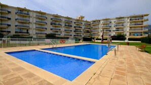 Un área de piscina con dos piscinas, rodeada de un solárium con baldosas y contigua a un edificio residencial de varios pisos.