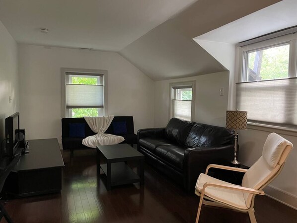 A living room with a black leather sofa, a white armchair, a coffee table, and a TV on a stand. There are two windows with blinds, and a lamp on a side table. - Fantastic updated apartment with parking (Glenside)
