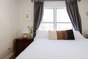 Una cama bien hecha con sábanas blancas y un cojín multicolores, una mesita de noche con una lámpara y una planta pequeña, y una ventana con cortinas grises.