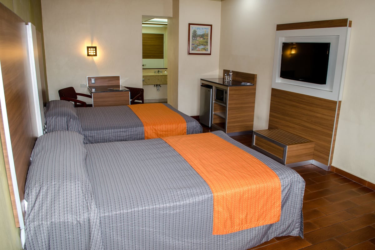 A hotel room with two beds, a desk, a TV, a mirror, and a bathroom visible in the background.