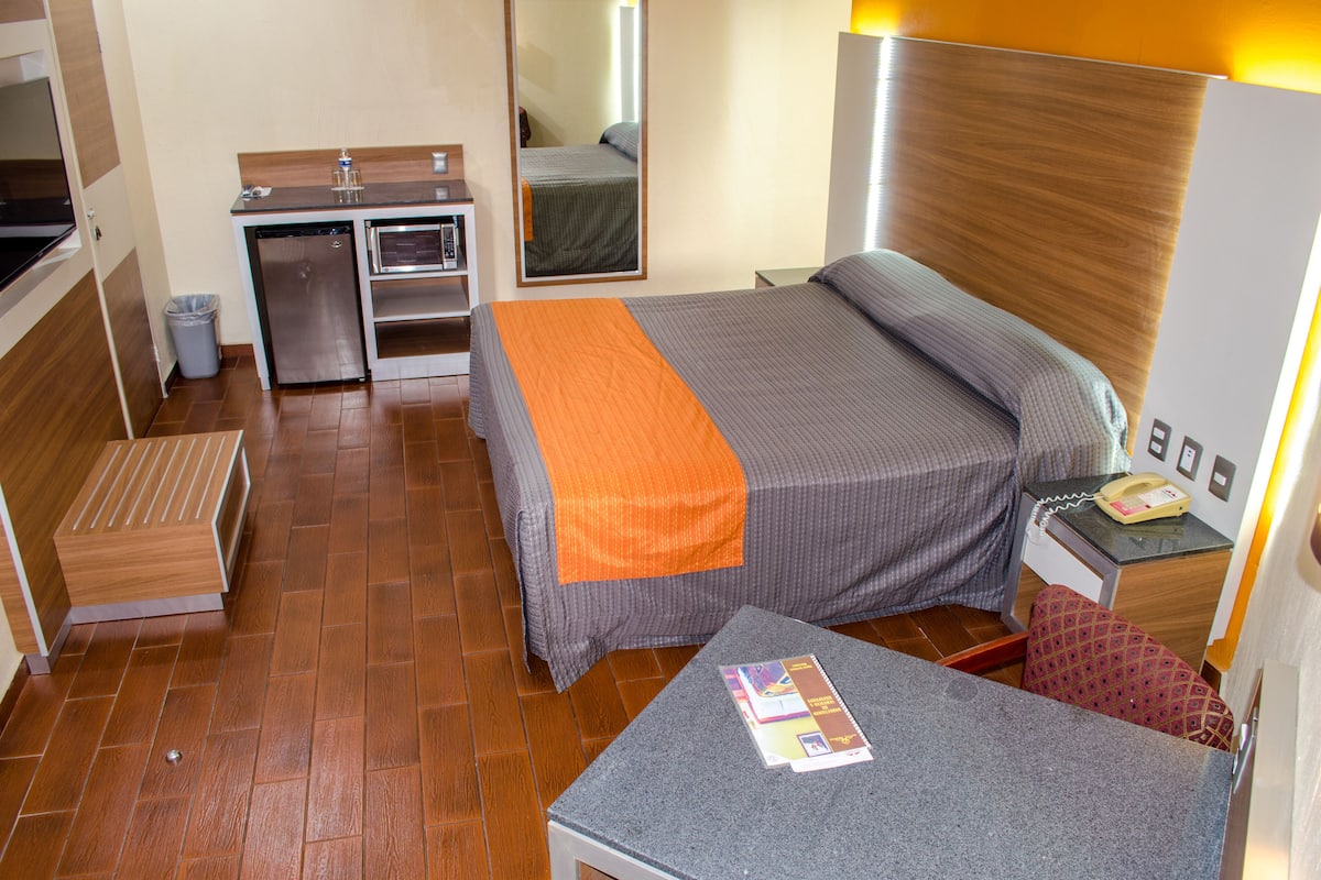 A compact hotel room with a bed, a small desk, a mirror, a bedside table with a phone, and a trash bin.