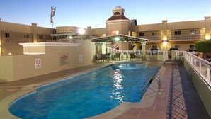 A swimming pool with a sunshade and lounge chairs, surrounded by a building with balconies and a tower. - Hotel Premier (Hermosillo)