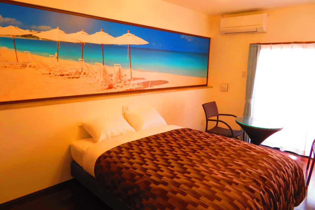 A hotel room with a bed, a chair, a painting of a beach scene, and an air conditioning unit.