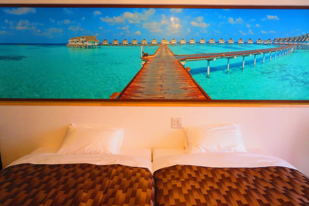 A hotel room with a bed featuring a woven headboard, a large window with a view of overwater bungalows and a wooden pier.