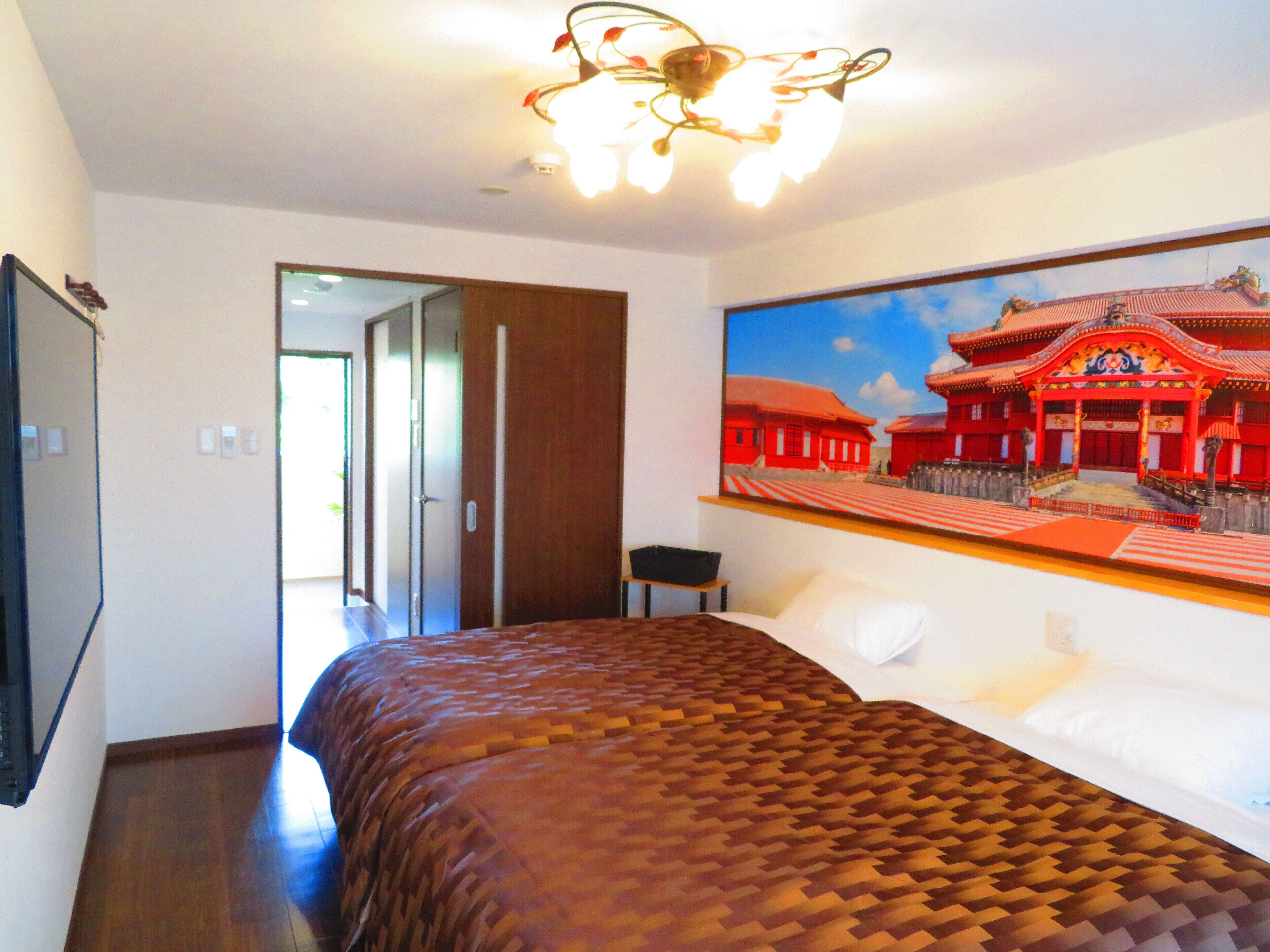 A hotel room with a bed, a painting of a traditional building, a ceiling light fixture, and a door leading to another room.